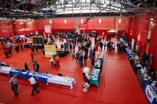 People walking around a large hall outlined by rows of tables.